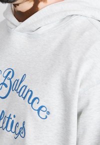 Primer plano de una sudadera ligera de color gris con el texto azul "New Balance Athletics" llevada por una persona con vitíligo visible en su cuello.
