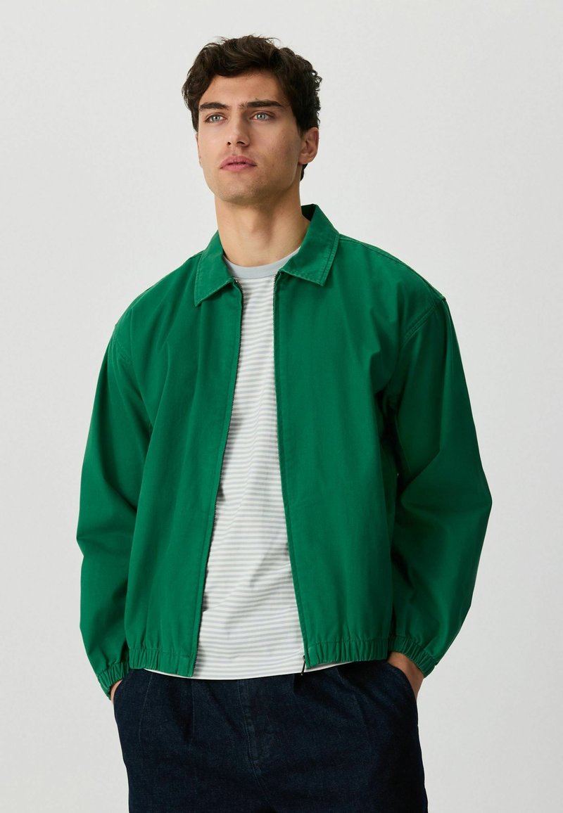 Jeune homme portant une veste verte à fermeture éclair sur un t-shirt blanc et gris à rayures, un pantalon foncé, les mains dans les poches, regardant devant lui.