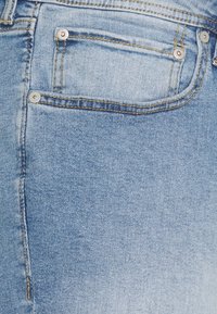 Svetle modrá džínovina s kapsou na zadní straně, viditelným šitím a kovovými nýtky. Textura vypadá jemně a s mírným vyblednutím.