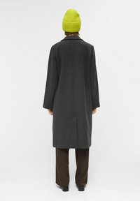 Abrigo de lana gris con silueta recta, mangas largas y una abertura en la parte posterior, acompañado de pantalones marrones y un gorro de punto verde brillante.
