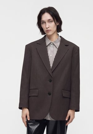 Personne aux cheveux courts et foncés portant un blazer sombre à larges rayures sur une chemise rayée boutonnée et un pantalon noir, debout devant un fond blanc.