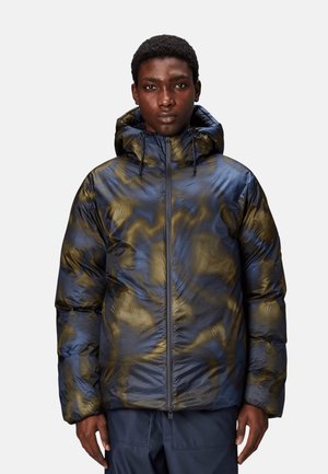 Modèle masculin portant une doudoune à capuche avec un motif camouflage bleu et olive et une fermeture éclair à l'avant, debout devant un fond uni.