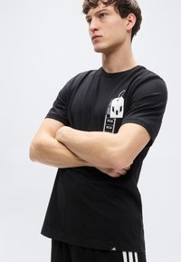 Μαύρο βαμβακερό t-shirt με κοντά μανίκια. Διαθέτει γραφική σχεδίαση στο αριστερό στήθος που απεικονίζει μια ετικέτα και έναν χαρακτήρα. Κανονική εφαρμογή.