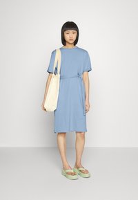 Vestido azul claro de mangas curtas com cintura ajustada, combinado com uma mala de mão creme e sandálias de plataforma verdes. Textura de tecido suave e casual.