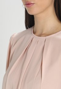Blusa rosa chiaro con design plissettato, scollatura rotonda e texture morbida. Il tessuto cade in modo fluido con una leggera lucentezza.