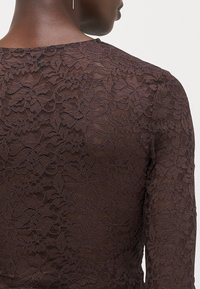 Brown lace top featuring a floral pattern, long sleeves, a rounded neckline, and a delicate, textured surface.