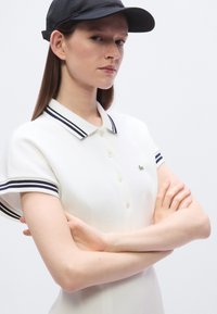 Polo blanc avec col et manches à rayures bleu marine, arborant un petit logo vert. Le modèle pose les bras croisés et porte une casquette noire.