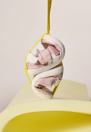 Zapatos pequeños de niño de color rosa y blanco con tiras de velcro colgando de cordones amarillos sobre una superficie curva de color amarillo pálido.