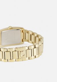 Montre en métal doré avec un boîtier rectangulaire, un bracelet en maillons et un fermoir circulaire. Surface polie et réfléchissante.