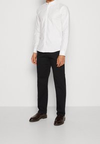 Witte langemouwen shirt met een Mandarin-kraag, gecombineerd met zwarte broek en donkerbruine schoenen. Het shirt heeft knoopsluitingen.