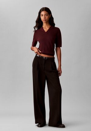 Femme aux longs cheveux foncés portant un pull bordeaux à manches courtes et un pantalon noir à jambes larges avec une ceinture sur un fond gris.