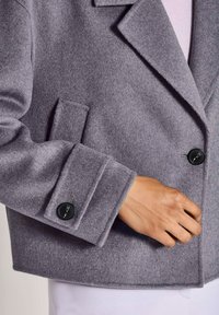 Grauer Wollmantel mit schwarzen Knöpfen, getragen über einem hellrosa Oberteil, wobei der Ärmelsaum und die Hand nahe der vorderen Kante des Mantels zu sehen sind.