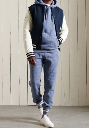 Mies, jolla on sininen huppari ja collegehousut, mustavalkoinen varsity-takki, valkoiset tennarit ja kuvioidut sukat, kävelee sisätiloissa puisella lattialla.