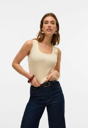 Débardeur beige côtelé avec un encolure carrée. Porté avec un jean taille haute bleu foncé et une ceinture noire. Présente un bracelet brun rouge.