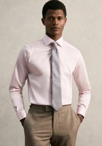Chemise rose pâle à col italien, associée à une cravate argentée à motifs et un pantalon sur mesure marron clair, avec une texture lisse.