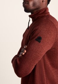 Red zip-up fleece sweater with a textured finish, featuring a high collar and a black logo patch on the left sleeve.