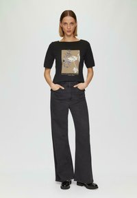Haut noir à manches courtes avec décolleté carré, ornée d'un motif floral en beige et bleu. Associé à un jean large foncé et des chaussures noires.