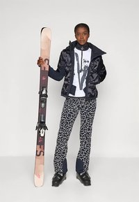 A sífelszerelés egy világos színű snowboardból áll fekete kötésekkel, sötét hőkabáttal, mintás nadrággal és síbakancsokkal.