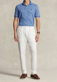 Man som bär en blå polo med segelbåtsmotiv, vita plisserade byxor, brunt läderklocka och bruna seglarskor med snörning.