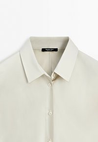 Světlá béžová košile na knoflíky s špičatým límečkem, třemi viditelnými knoflíky a černou etiketou uvnitř límečku s nápisem "Massimo Dutti."