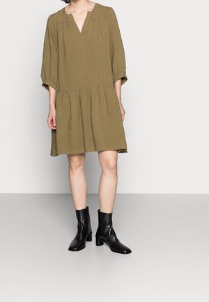 Kvinde iført en olivengrøn knælængde kjole med 3/4 ærmer og sorte læderankelstøvler, stående foran en ensfarvet baggrund.