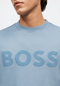 Άντρας που φοράει ανοιχτό μπλε φούτερ με ανεβασμένα γράμματα που σχηματίζουν τη λέξη «BOSS» στο στήθος, το οποίο κόβεται κάτω από τη μύτη.