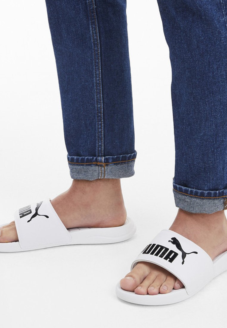 Sandales blanches avec une large lanière arborant le texte et le logo noir "PUMA". Portées avec un jean en denim bleu coupé, montrant des ourlets retroussés.