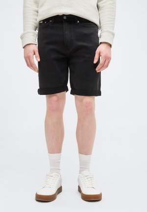 Person wearing black rolled-up denim shorts, beige sweatshirt, white socks, and white sneakers with brown soles, standing against a plain background.