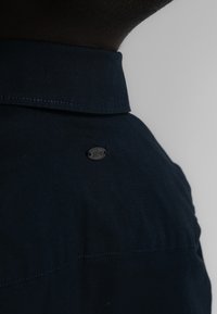 Navyblauwe geknoopte shirt met een gladde textuur en subtiele stiksels, met een klein donker logo label op de achterkant nabij de kraag.