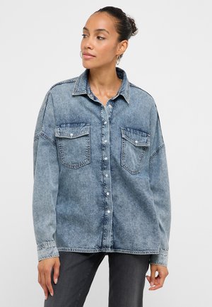 Frau mit zusammengebundenem Haar zu einem Dutt, die Creolen trägt und ein hellblaues Jeanshemd mit zwei Vordertaschen sowie dunkle Hose anhat.