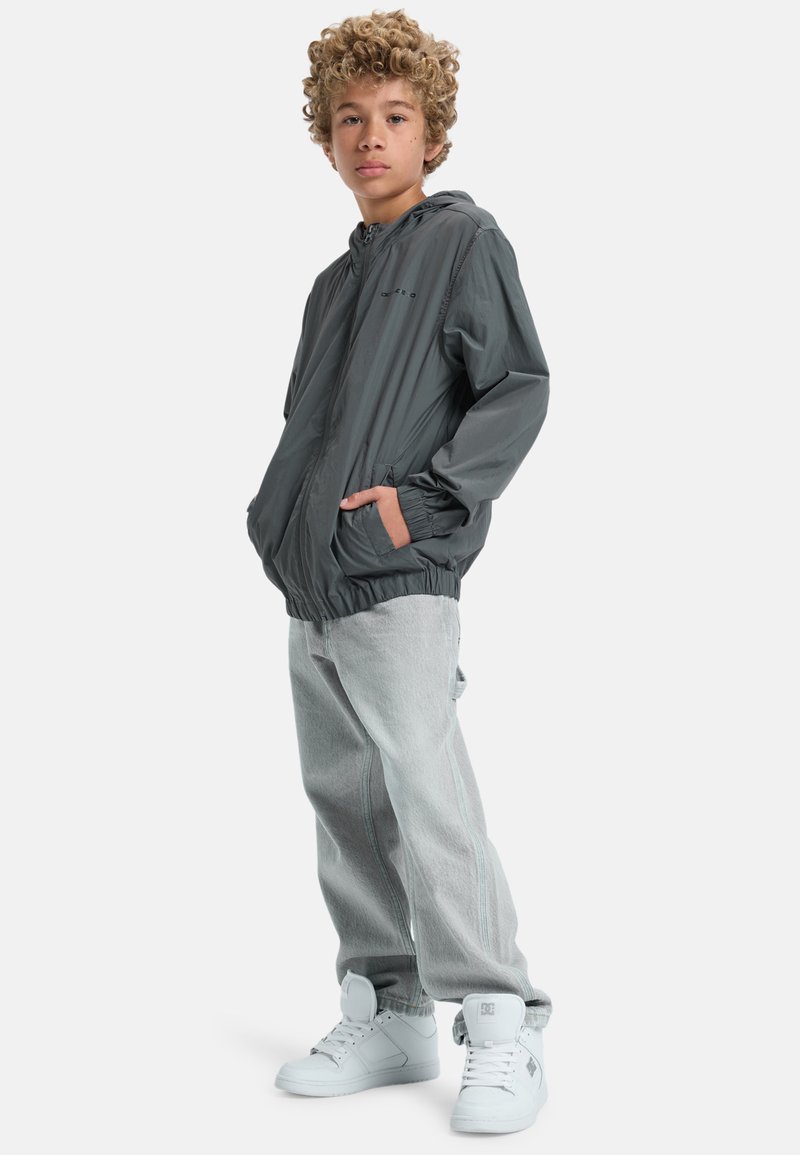 Grå lättviktsjacka med huood och elastiska ärmar, paired med ljusgrå jeans och vita högskor. Enkelt design, inga mönster.