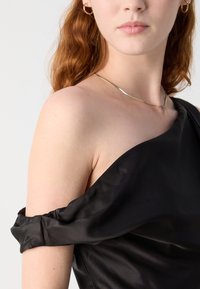 Jeune femme aux cheveux roux portant une robe en satin noir à épaules dénudées, un collier en or et des boucles d'oreilles créoles en or sur un fond blanc.