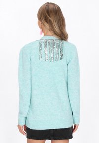 Világoskék, bolyhos pulóver hosszú ujjakkal, ezüst rojt díszítéssel a háton és bordázott szegéllyel.