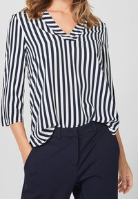 Blusa a righe blu navy e bianche con scollatura a V e maniche a tre quarti, realizzata in tessuto leggero, con un dettaglio di tasca frontale.