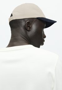 Beige cap with a navy peak, constructed from fabric, featuring a textured finish and an adjustable strap at the back. White shirt visible below.
