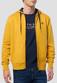 Uomo che indossa una felpa con zip giallo senape sopra una t-shirt blu navy con disegni di bandiere nautiche e jeans blu, su uno sfondo grigio semplice.