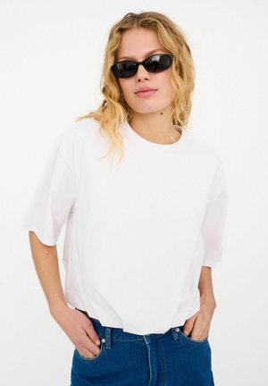White oversized short-sleeve t-shirt with a round neckline, paired with blue denim jeans. The model wears black sunglasses.