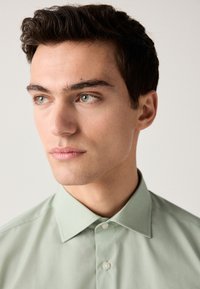 Lysegrøn skjorte med krave og en glat tekstur, der har lukning med knapper og subtile syningsdetaljer, båret af en ung mand med kort hår.