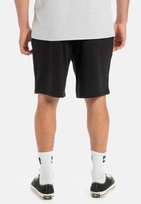 Pantalones cortos de algodón negros con un ajuste relajado, que cuentan con un bolsillo trasero, combinados con calcetines blancos y zapatillas negras. Camiseta gris visible en la parte superior.
