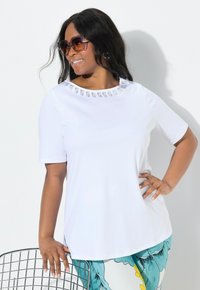 Camiseta blanca de manga corta con un cuello redondo que presenta acentos decorativos. La modelo lleva gafas de sol y está de pie al lado de una silla de alambre.