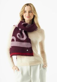 Wijnrood en lichtpaars sjaal met patroon en rafelige randen, met een klein groen logo; gedragen over een trui in neutrale tinten.