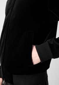 Черен велурен яке с цип, с Ribbed маншети, странични джобове и гладка текстура, представящо елегантен и модерен дизайн.
