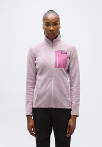 Γυναίκα που φοράει μωβ ανοιχτό μοτίβο φλις ζακέτα με ροζ τσέπη στο στήθος, στέκεται μπροστά σε απλό λευκό φόντο.