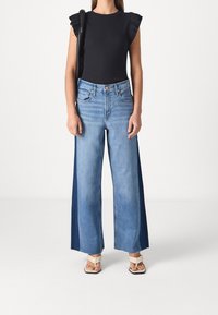 Svart ärmlös topp med volangärmar, matchad med vida blå jeans med tvåtonskomposition, avslutad med beiga sandaler.