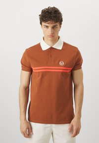 Ruda polo marškinėliai su balta apykakle, turintys horizontalių rožinių juostelių ir nedidelį logotipą kairėje krūtinės pusėje. Trumpomis rankovėmis, pagaminti iš medvilnės.