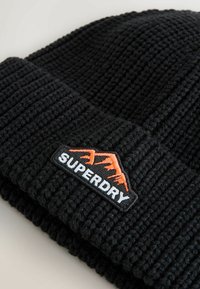 Zwarte gebreide beanie met ribbeltextuur, met een opvallende patch waarop het woord "SUPERDRY" en een bergafbeelding in oranje staan.
