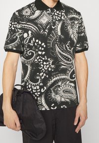 Czarny i biały polo w wzór paisley z krótkimi rękawami i czarnym kołnierzykiem, trzymany w poziomej orientacji razem z czarną torbą.