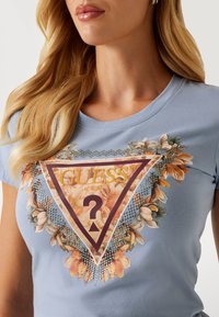 Gaiši zils pieguļošs T-krekls ar ziedu grafiku, trīsstūrveida "GUESS" logo un sudraba rotājumiem ap dizainu ar teksturētu apdari.