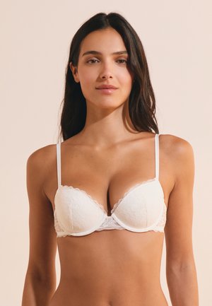 Mujer con cabello largo y oscuro, vistiendo un sujetador blanco de encaje, mirando hacia adelante frente a un fondo beige liso.