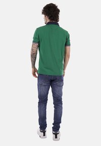 Homme tatoué portant un polo vert à manches courtes, un jean bleu et des baskets blanches, debout de dos sur un fond uni.